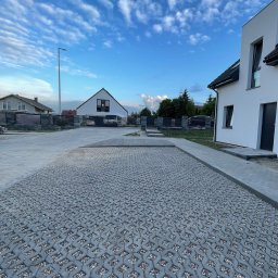 Metrikum Aleksander Atraszkiewicz - Szara, ażurowa kostka betonowa wypełniona drobnym kamieniem na podjeździe przed nowoczesnym domem. Błękitne niebo z chmurami w tle.