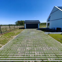 Metrikum Aleksander Atraszkiewicz - Nowoczesny podjazd z ażurowej kostki betonowej, porośniętej trawą, prowadzący do garażu i domu w minimalistycznym stylu. Błękitne niebo w tle.