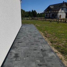 Metrikum Aleksander Atraszkiewicz - Ścieżka z szarej kostki brukowej prowadząca wzdłuż białego budynku, z widokiem na trawnik i dom w tle, w słoneczny dzień. Różne rozmiary kostek tworzą unikalny wzór.