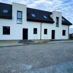 Nowoczesne bliźniaki z białymi elewacjami, czarnymi dachami i oknami, oraz podjazdem wyłożonym ażurową kostką betonową na tle błękitnego nieba z chmurami.