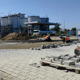 Dwóch pracowników w kamizelkach odblaskowych układa szarą kostkę brukową na placu przemysłowym z widocznymi zbiornikami i maszyną do cięcia kostki.