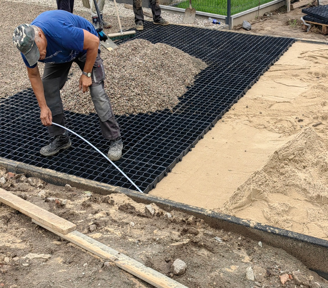 Pracownik układa czarną, plastikową kratkę drenażową na podsypce z piasku i żwiru, przygotowując podłoże pod nawierzchnię. Widoczne narzędzia i materiały budowlane.