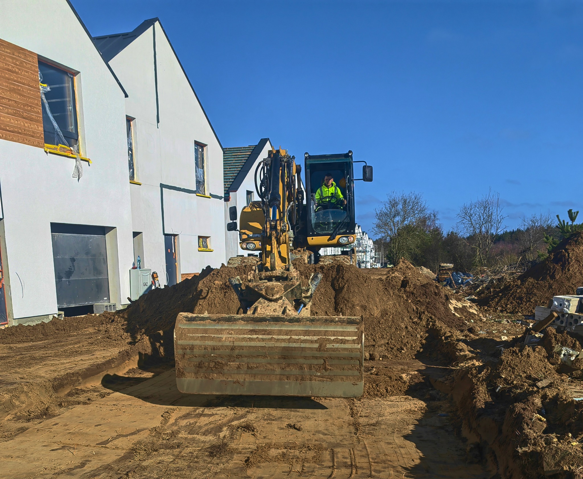Żółta koparka Caterpillar z operatorem w jaskrawej kamizelce, przygotowująca teren pod budowę domu jednorodzinnego z garażem i elementami drewnianej elewacji.