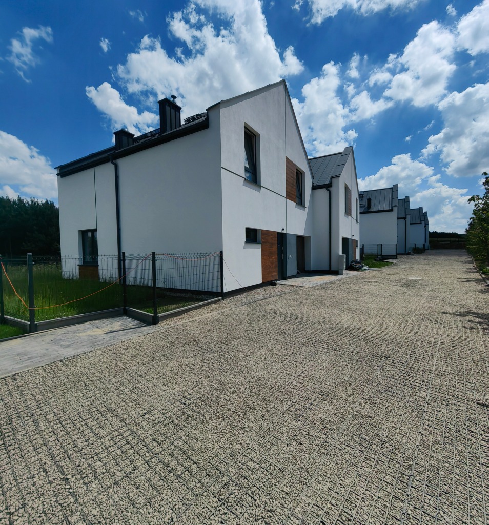 Nowoczesne szeregowe domy z jasną elewacją i ciemnymi dachami, wykończone z użyciem gabionów wypełnionych jasnym kamieniem, pod błękitnym niebem z białymi chmurami.