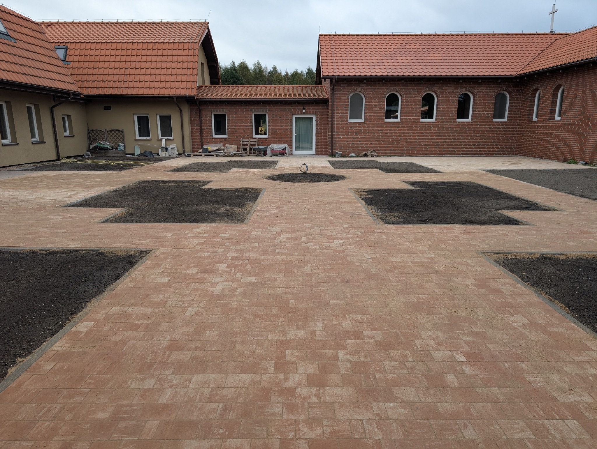 Ceglana nawierzchnia z kwadratowymi rabatami ziemi na dziedzińcu, otoczona budynkami z czerwonej cegły i dachówką. Widok z góry na symetryczny wzór.