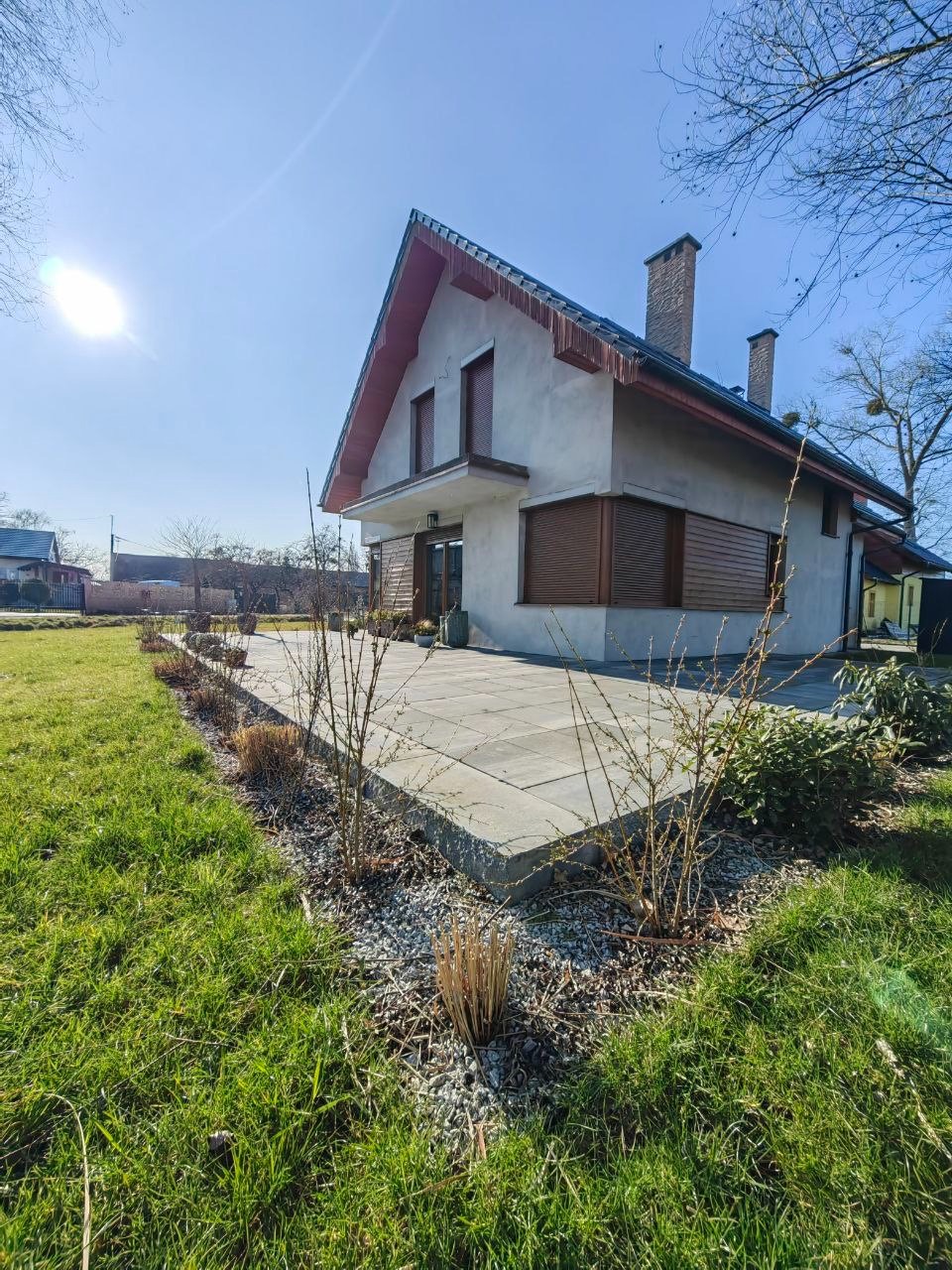 Nowoczesny taras z szarej kostki betonowej przy domu jednorodzinnym, z widocznym trawnikiem i elementami zieleni. Słoneczny dzień.