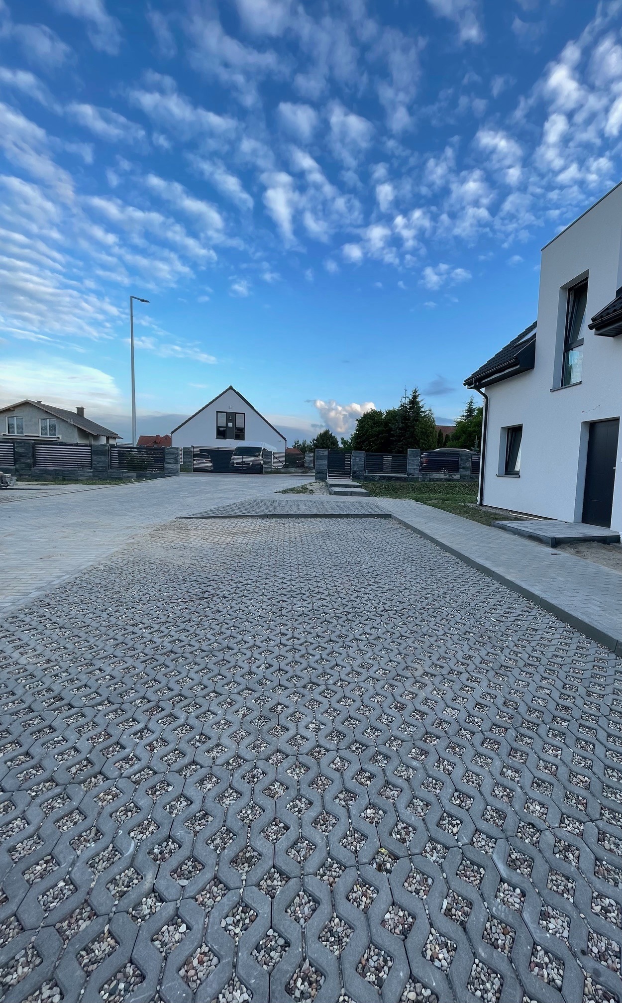Szara, ażurowa kostka betonowa wypełniona drobnym kamieniem na podjeździe przed nowoczesnym domem. Błękitne niebo z chmurami w tle.