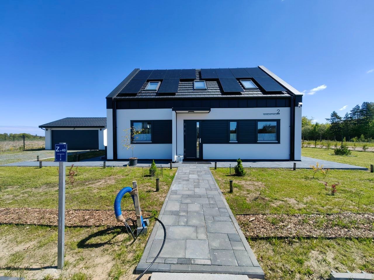 Nowoczesny dom jednorodzinny z czarnym dachem i panelami słonecznymi, do którego prowadzi ścieżka z szarej kostki brukowej. Widoczny garaż i zadbany trawnik.