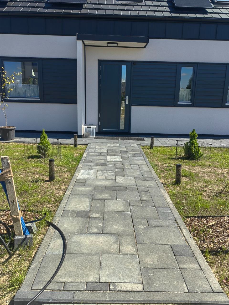 Nowoczesny chodnik z szarej kostki betonowej prowadzący do domu z ciemną stolarką okienną i drzwiami. Uzupełnieniem są małe drzewka i drewniane paliki.