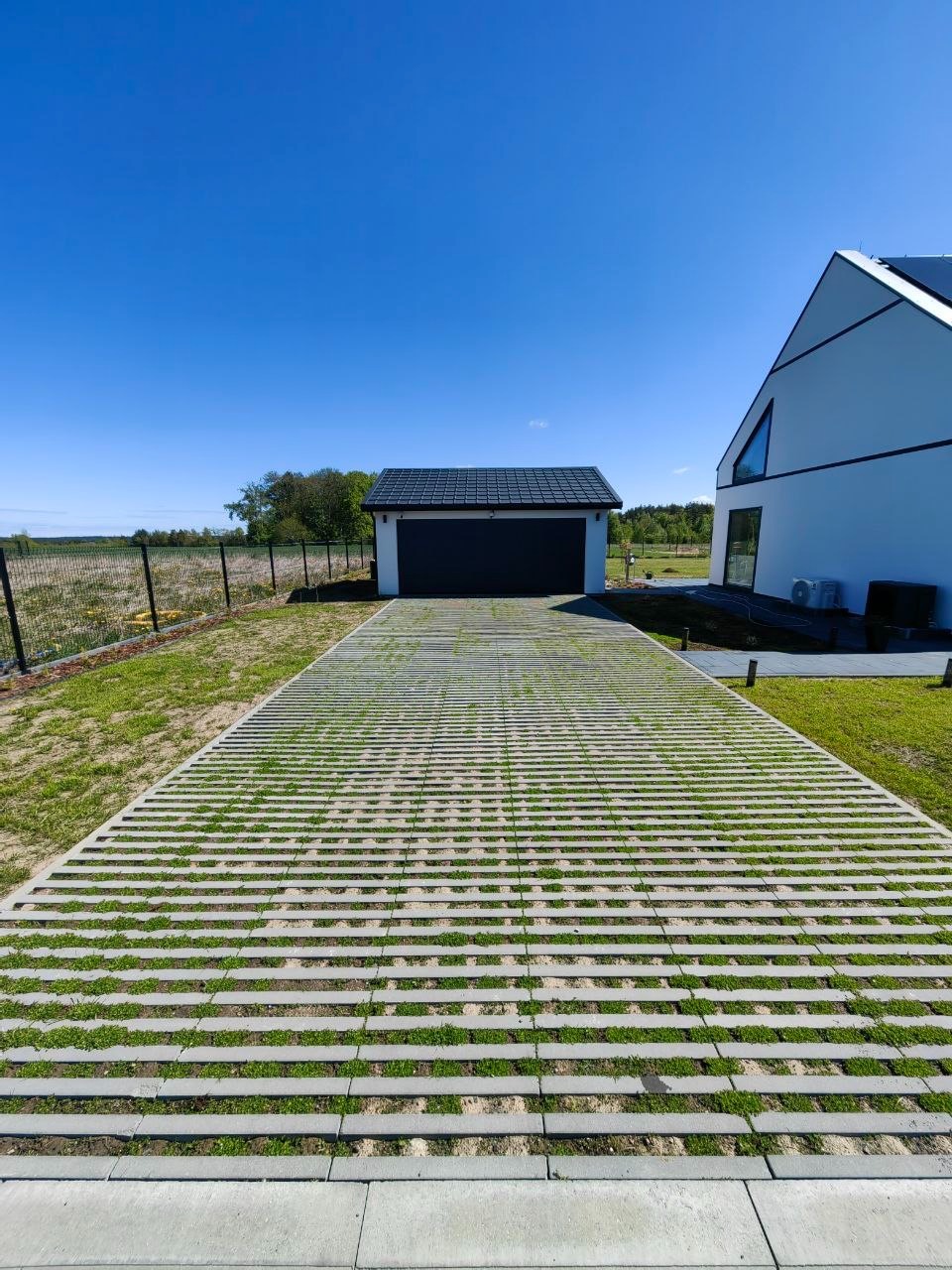 Nowoczesny podjazd z ażurowej kostki betonowej, porośniętej trawą, prowadzący do garażu i domu w minimalistycznym stylu. Błękitne niebo w tle.
