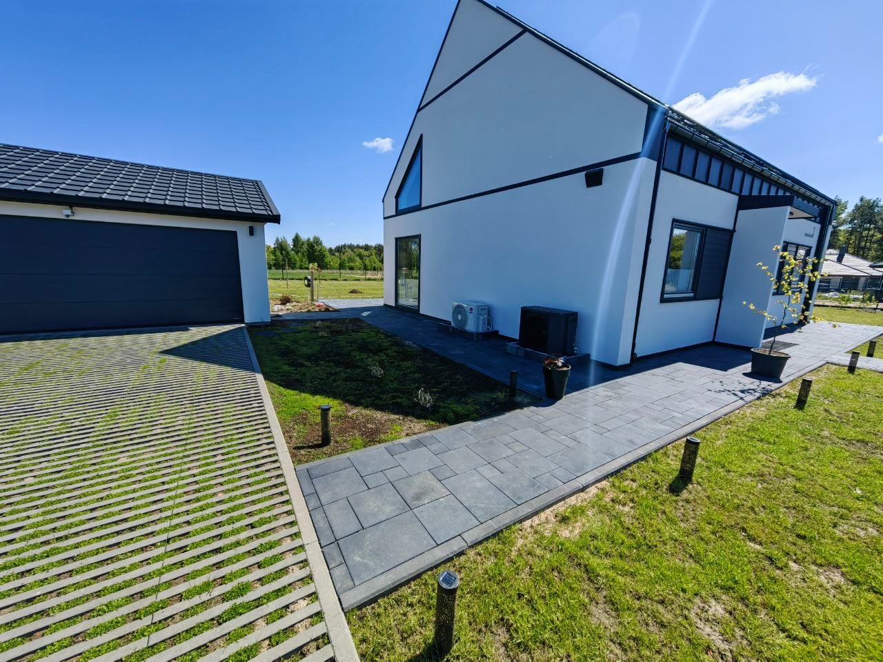 Nowoczesny dom z szarą ścieżką z płyt i ażurowym podjazdem porośniętym trawą. Minimalistyczny design, błękitne niebo, zadbany trawnik i oświetlenie zewnętrzne.