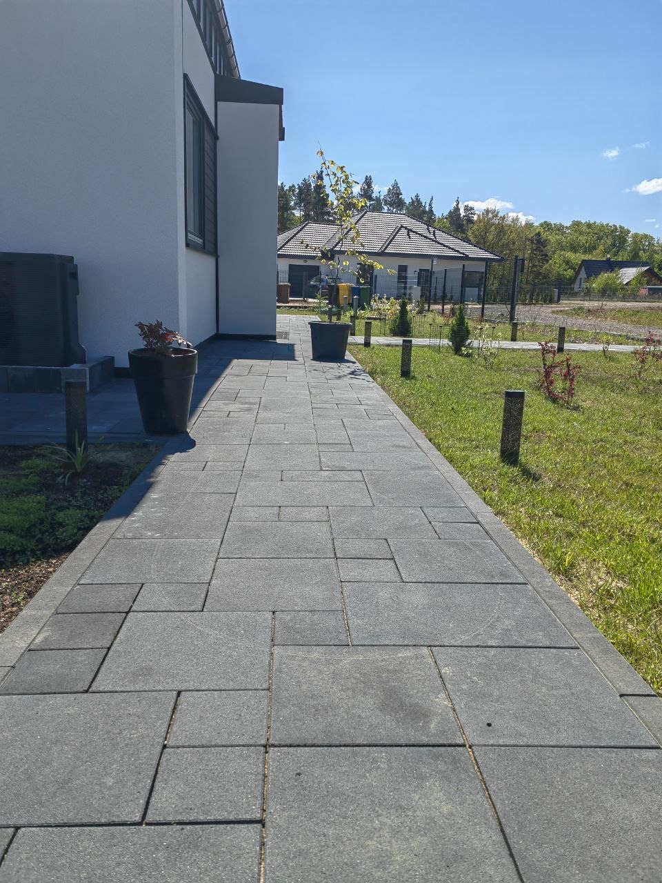 Nowoczesna ścieżka z szarej kostki betonowej prowadząca do domu, z donicami i młodymi drzewkami po bokach. Słoneczny dzień, widok na dom w tle.