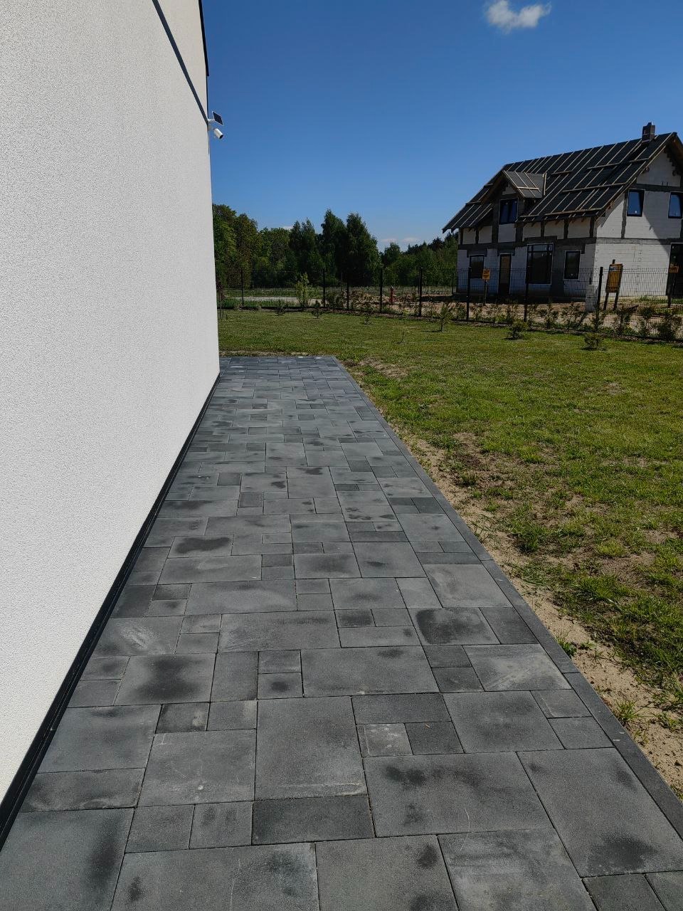 Ścieżka z szarej kostki brukowej prowadząca wzdłuż białego budynku, z widokiem na trawnik i dom w tle, w słoneczny dzień. Różne rozmiary kostek tworzą unikalny wzór.