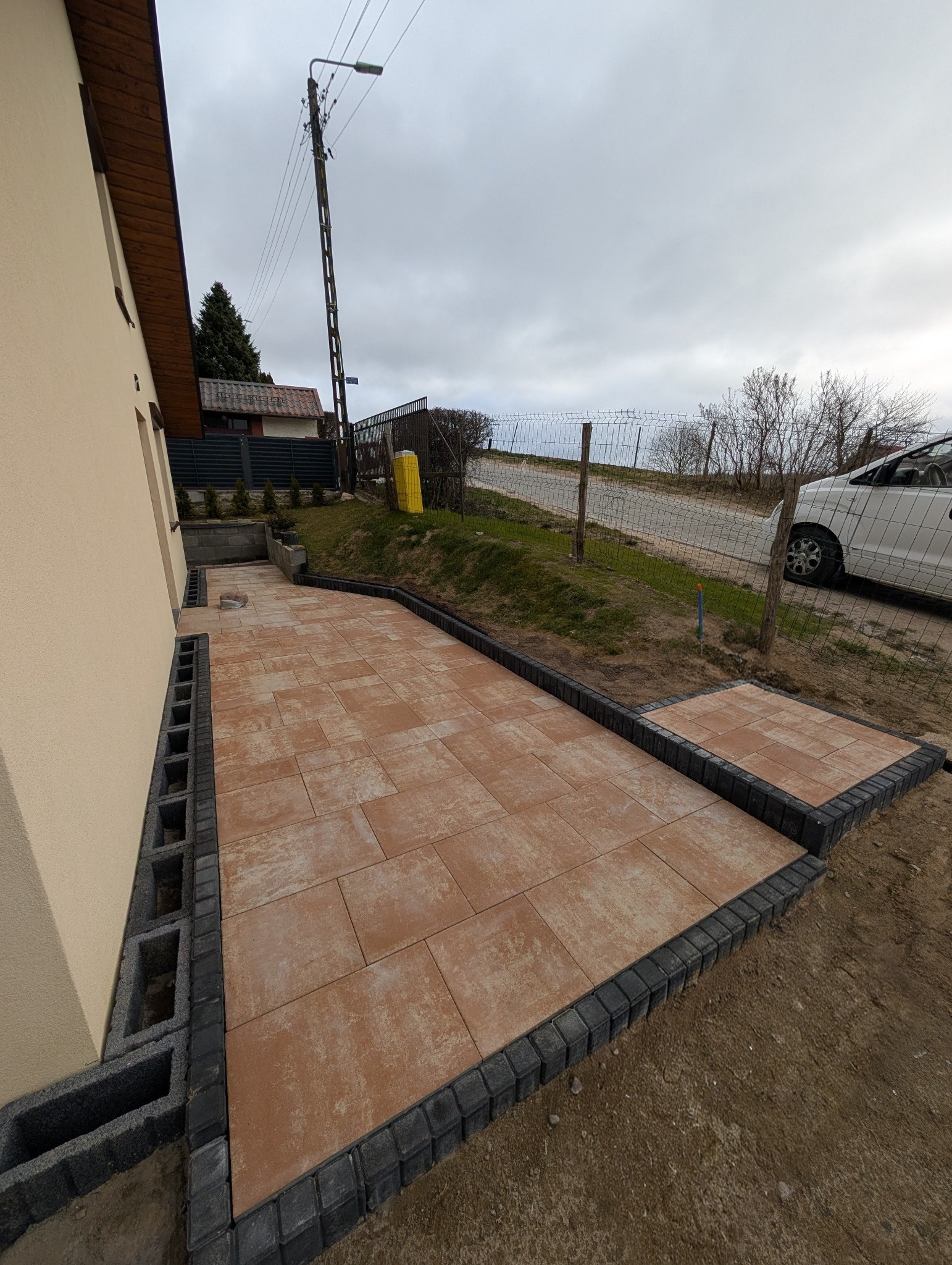 Nowy chodnik z brązowej kostki, obrzeża z szarej kostki, widok z góry, dom z beżową elewacją, słup oświetleniowy w tle, pochmurne niebo.