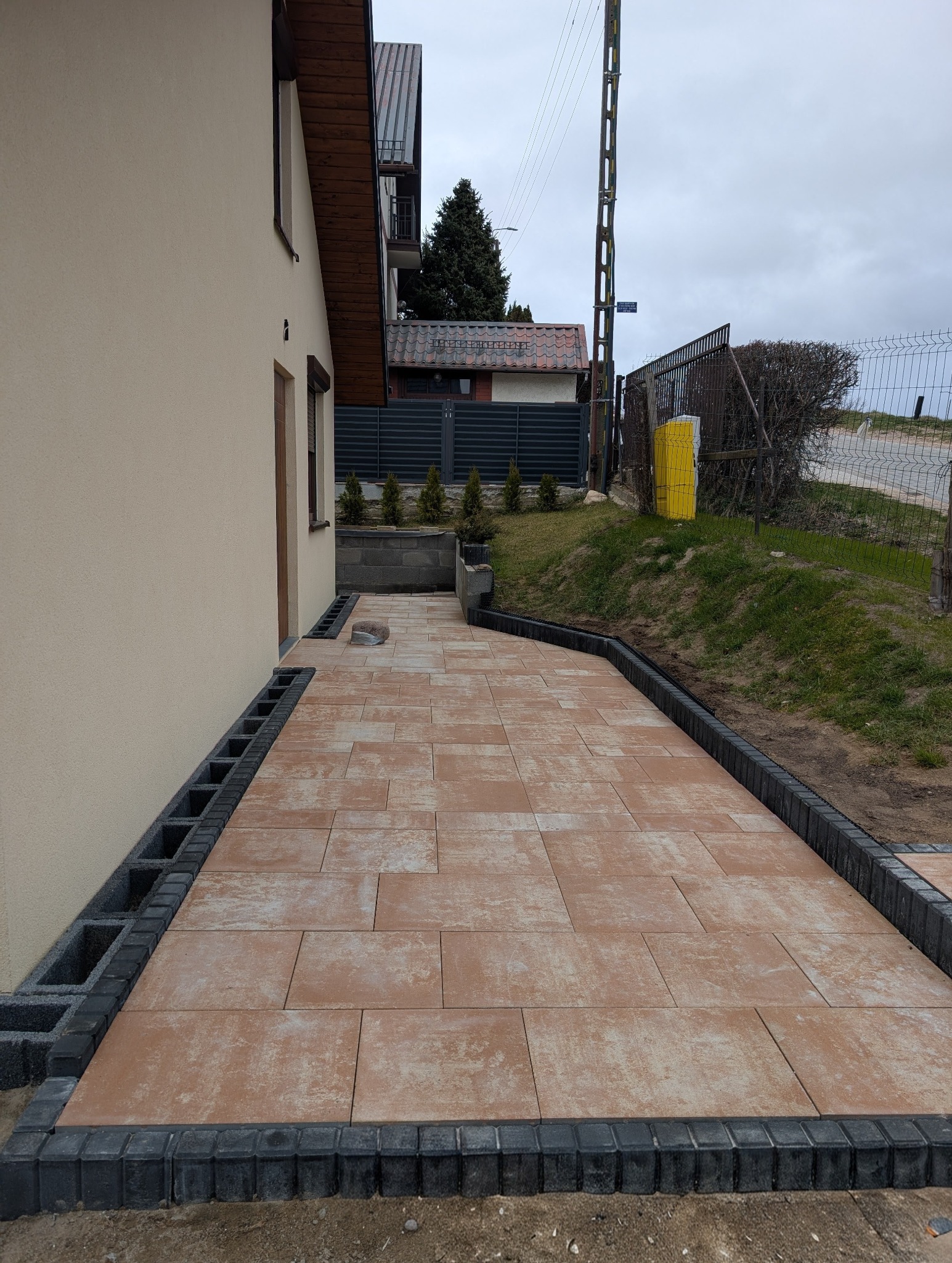 Ułożona kostka brukowa w odcieniach brązu, obramowana czarnym obrzeżem, przylegająca do ściany budynku. Widoczne elementy wykończeniowe i trawnik w tle.