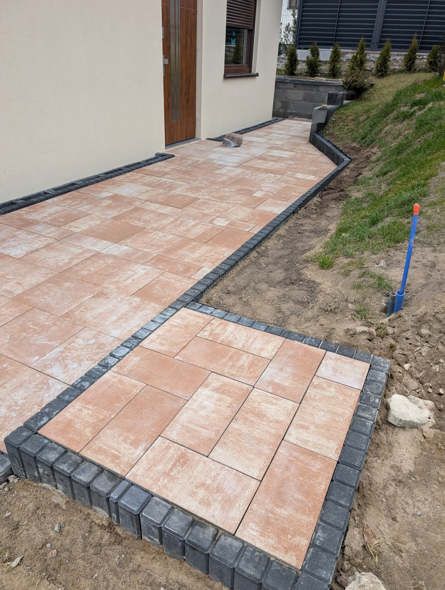 Nowa, beżowo-brązowa nawierzchnia z kostki betonowej, obramowana ciemnoszarą kostką, prowadząca do drzwi domu. Widoczny fragment trawnika i elewacji.