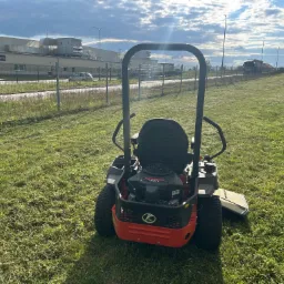 Czerwona kosiarka samojezdna na trawniku po koszeniu, z widocznymi śladami cięcia. W tle budynek przemysłowy i pochmurne niebo.