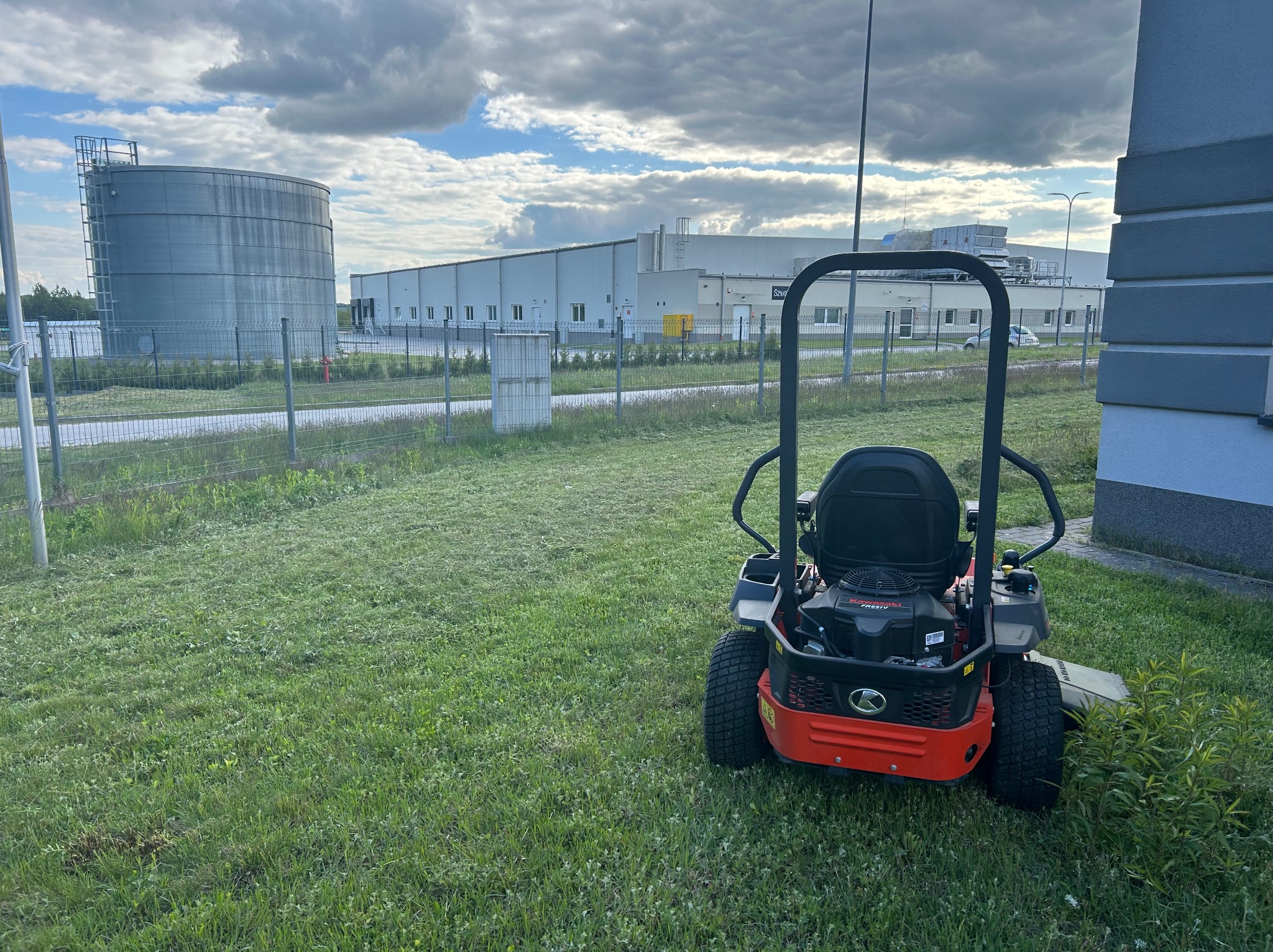 Czerwona kosiarka samojezdna na tle skoszonego trawnika przed budynkiem przemysłowym i zbiornikiem. Widoczne ślady koszenia.
