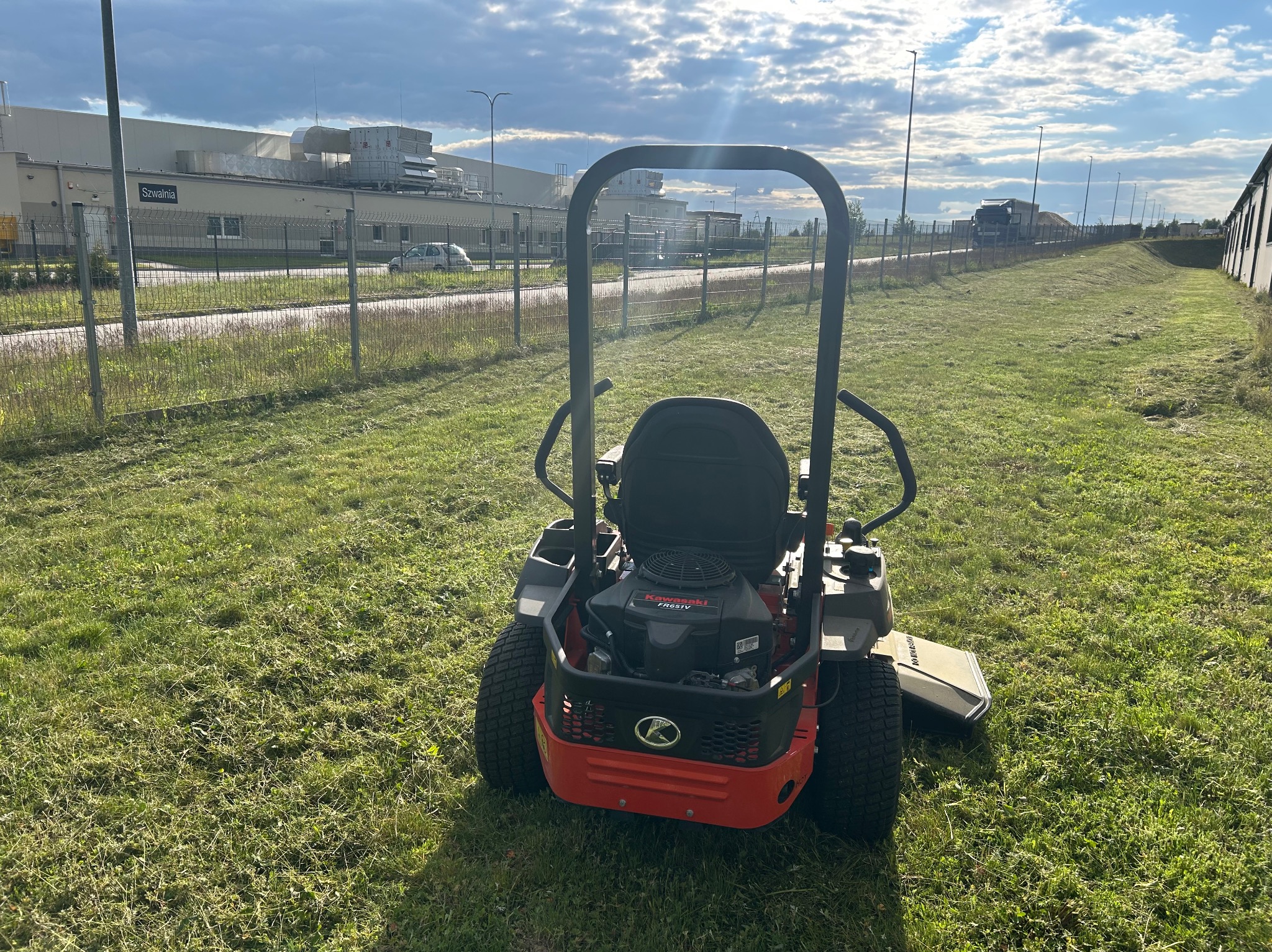 Czerwona kosiarka samojezdna na trawniku po koszeniu, z widocznymi śladami cięcia. W tle budynek przemysłowy i pochmurne niebo.