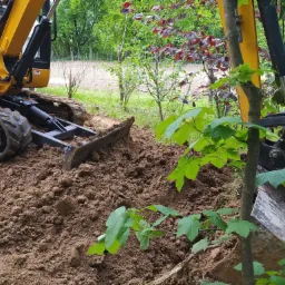 Żółta minikoparka z lemieszem częściowo zanurzona w brązowej ziemi, przygotowująca teren pod budowę, z zielenią w tle.