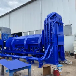 Pomalowana na niebiesko maszyna przemysłowa, z widocznymi przewodami hydraulicznymi i elementami konstrukcyjnymi, stojąca na zewnątrz budynku przemysłowego w pochmurny dzień.