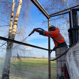 Kakadu - Pracownik w pomarańczowej kurtce, na podnośniku koszowym, czyści strumieniem wody konstrukcję stalową na tle drzew i pola.