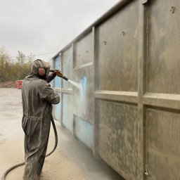 Kakadu - Pracownik w kombinezonie ochronnym piaskuje metalowy kontener na zewnątrz, widoczne ślady niebieskiej farby pod usuwaną warstwą.