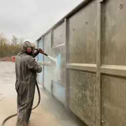 Pracownik w kombinezonie ochronnym piaskuje metalowy kontener na zewnątrz, widoczne ślady niebieskiej farby pod usuwaną warstwą.