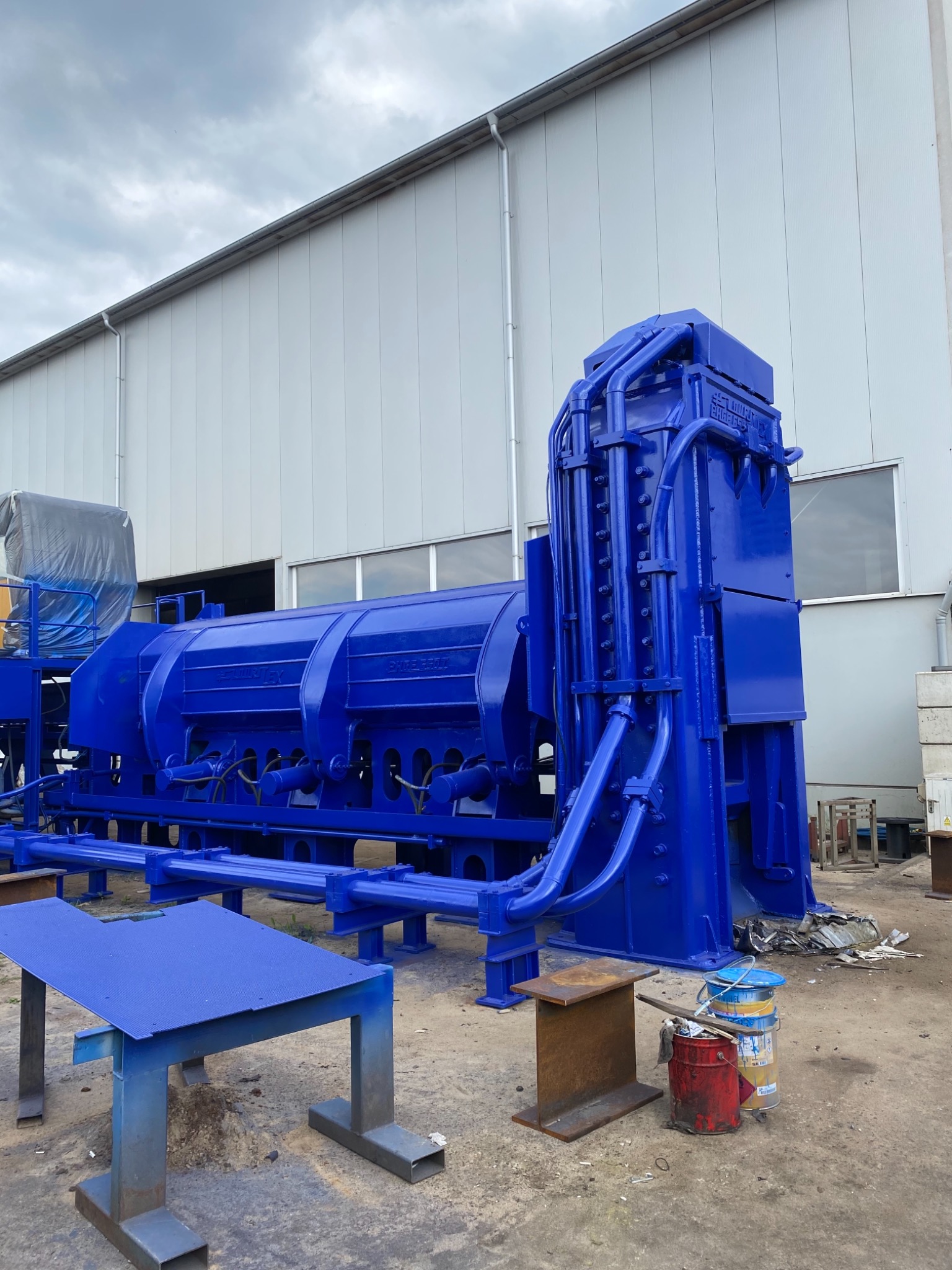 Pomalowana na niebiesko maszyna przemysłowa, z widocznymi przewodami hydraulicznymi i elementami konstrukcyjnymi, stojąca na zewnątrz budynku przemysłowego w pochmurny dzień.