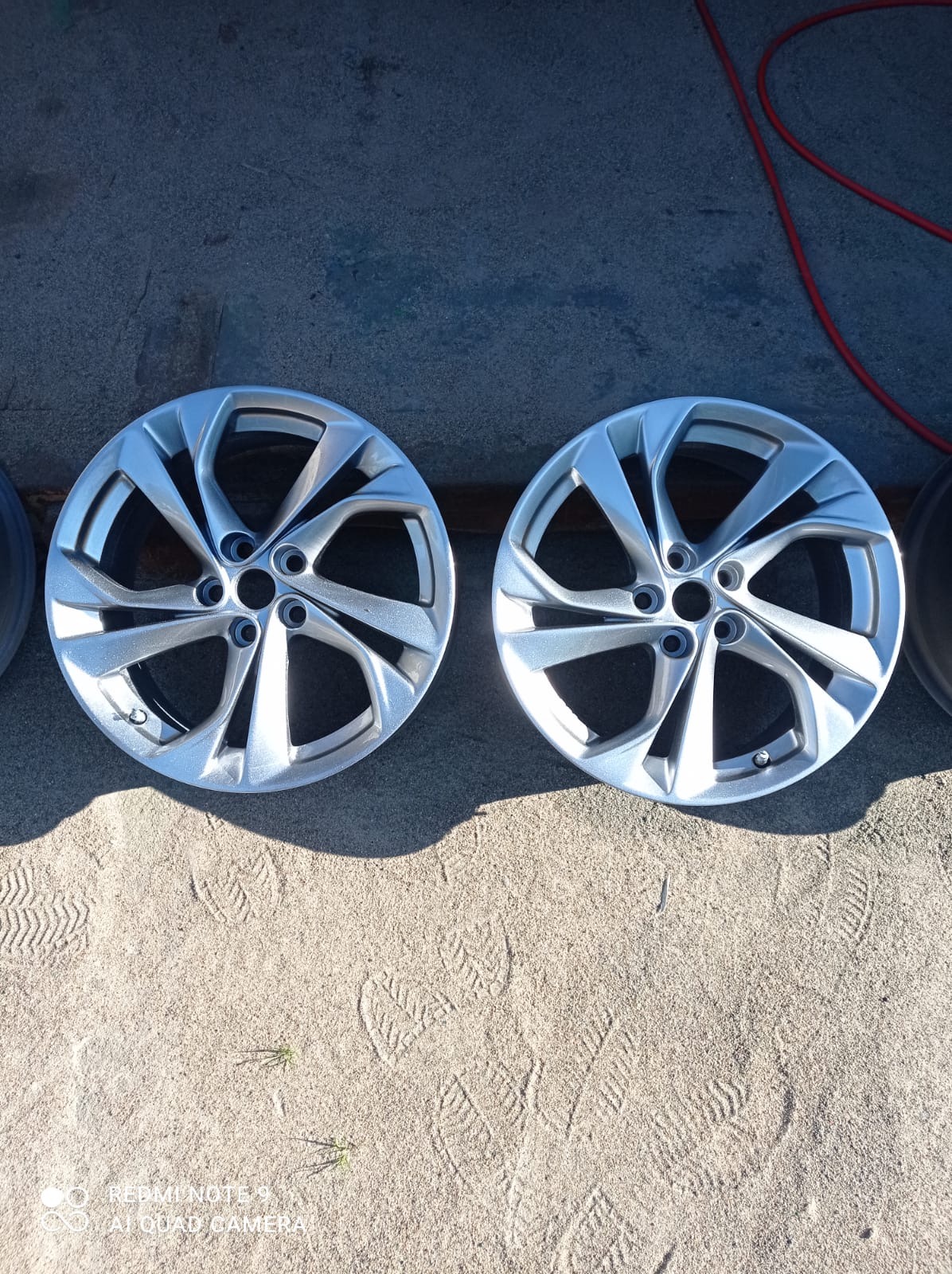 Dwa srebrne felgi aluminiowe z wieloramiennym wzorem leżą na piasku z odciskami stóp, obok widoczny fragment czerwonego węża.