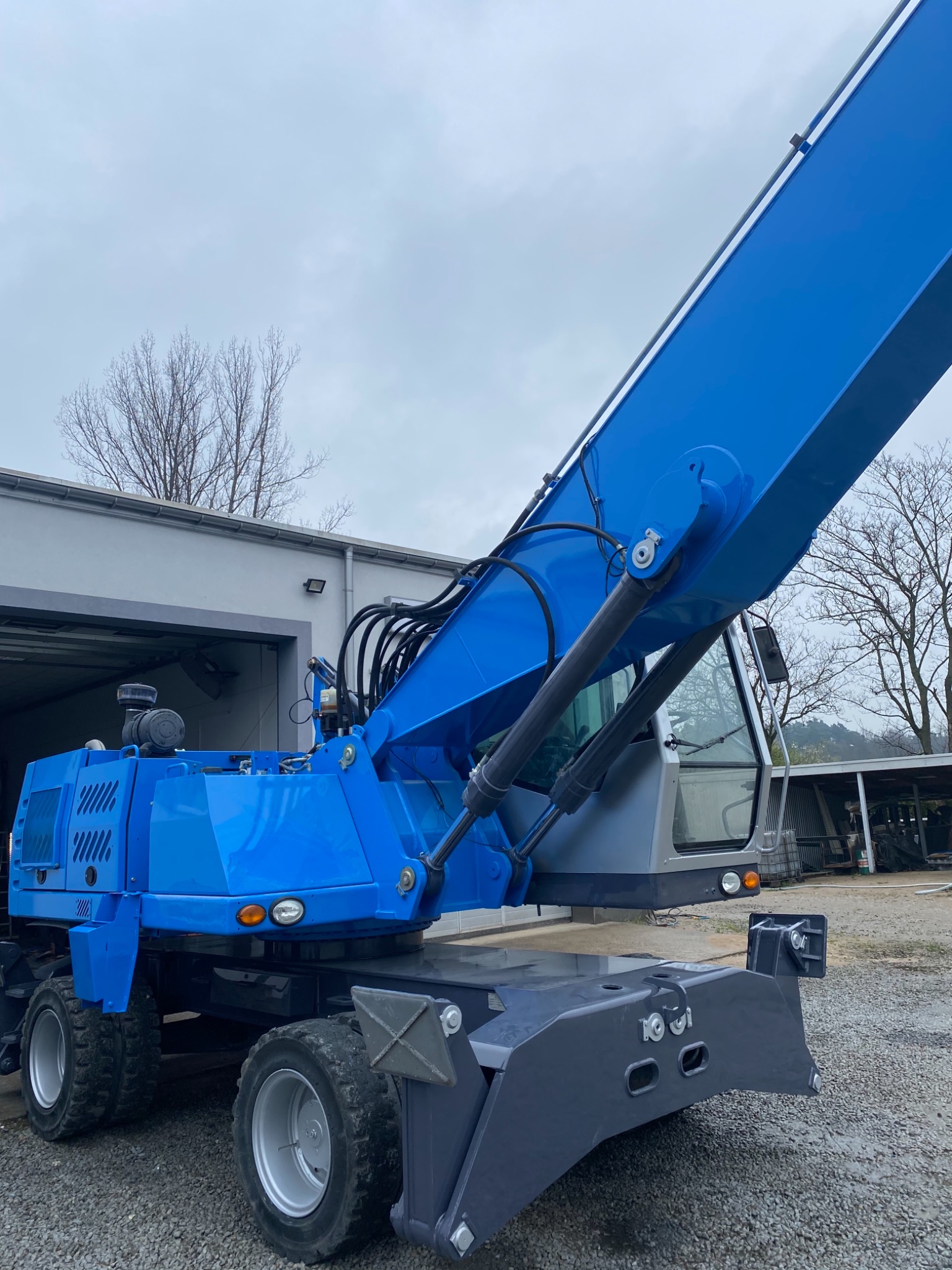 Niebieski, kołowy dźwig budowlany z wysięgnikiem, stojący na żwirowym placu przed budynkiem gospodarczym w pochmurny dzień.