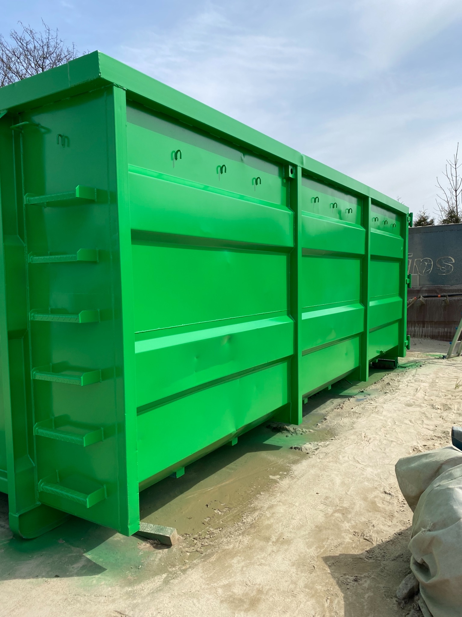 Duży, metalowy kontener na gruz w jaskrawo zielonym kolorze, świeżo po malowaniu, stojący na piaszczystym podłożu.