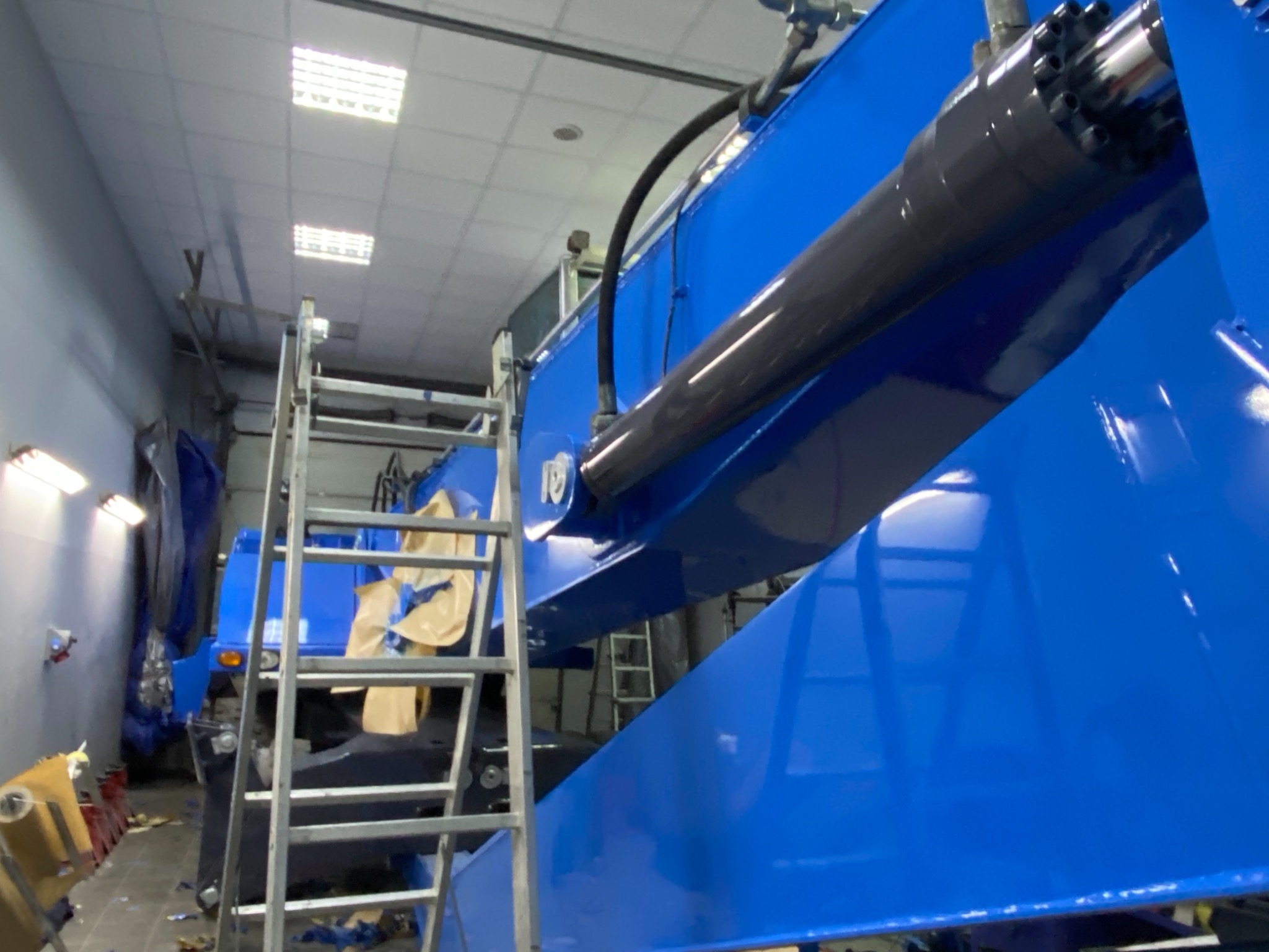 Niebieska konstrukcja stalowa z siłownikiem hydraulicznym, widoczna drabina i oświetlenie warsztatowe.