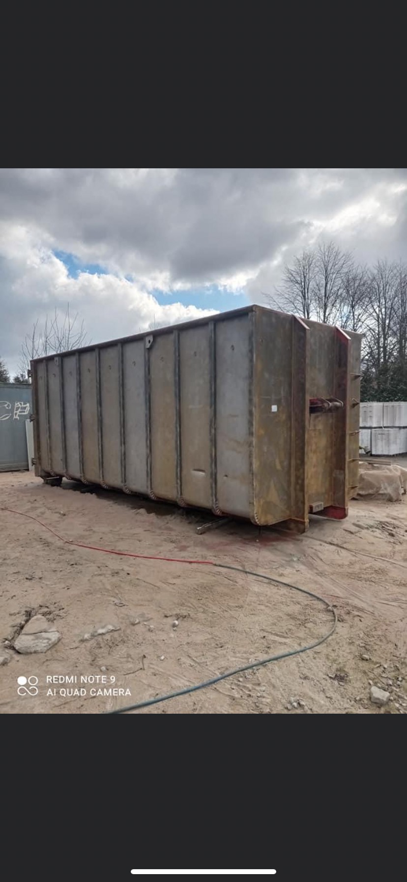 Duży, stalowy kontener hakowy na piasku, widoczne ślady korozji i niedokładnego czyszczenia, przewody leżące na ziemi.