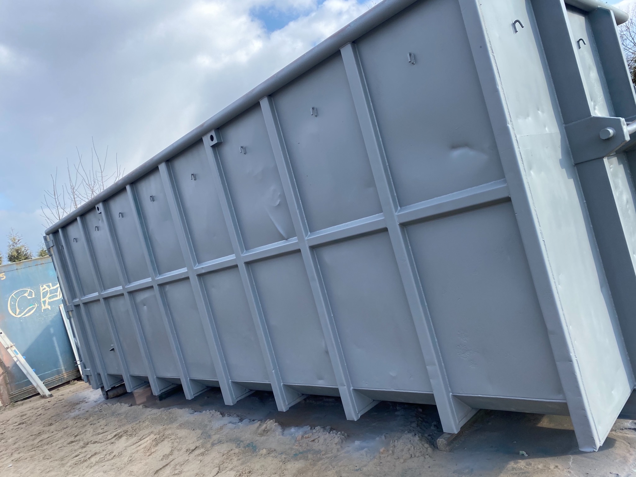 Duży, szary kontener z widocznymi śladami spawania i niedoskonałościami na powierzchni, stojący na piasku, z fragmentem drugiego kontenera w tle.