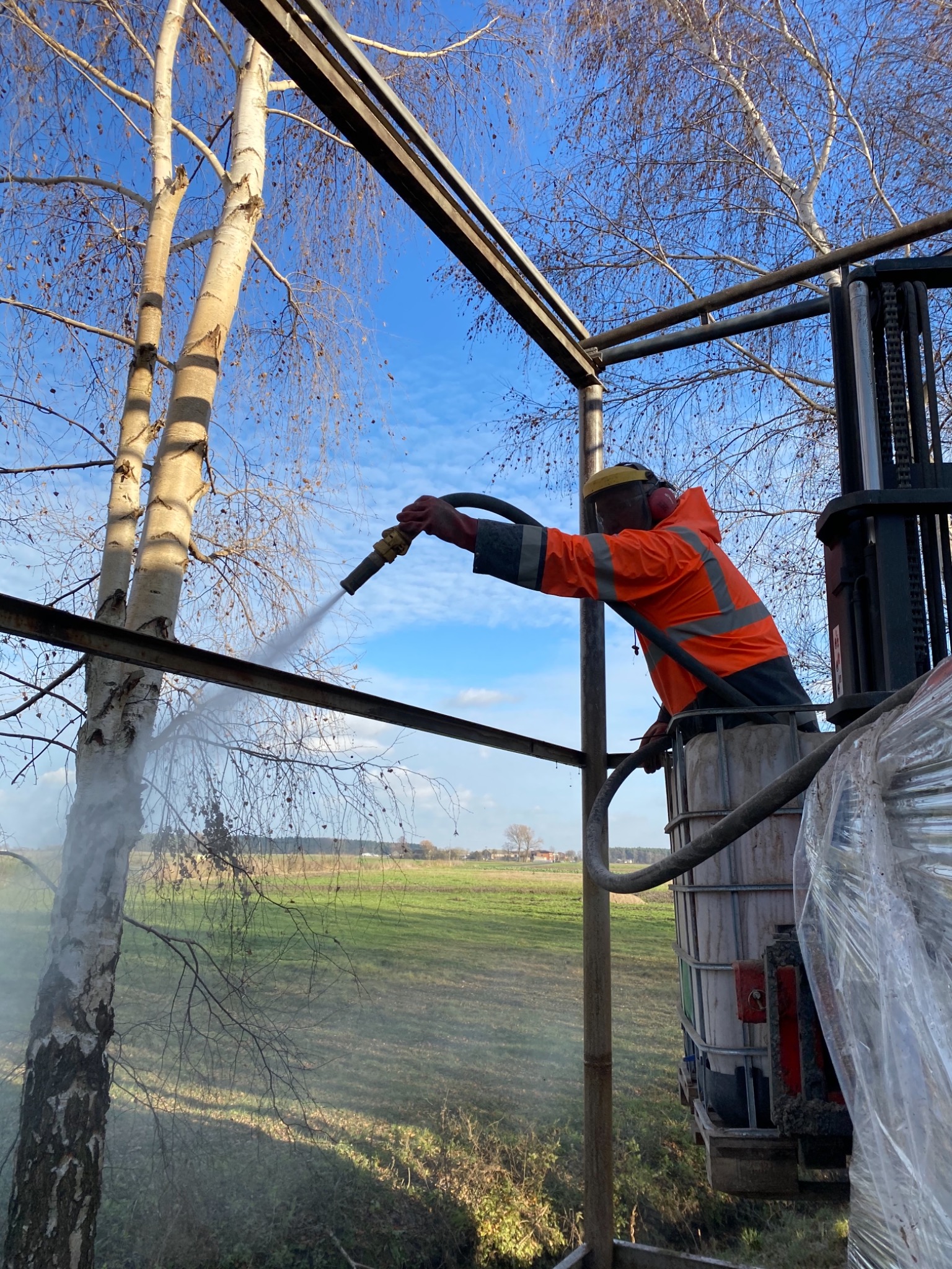 Pracownik w pomarańczowej kurtce, na podnośniku koszowym, czyści strumieniem wody konstrukcję stalową na tle drzew i pola.