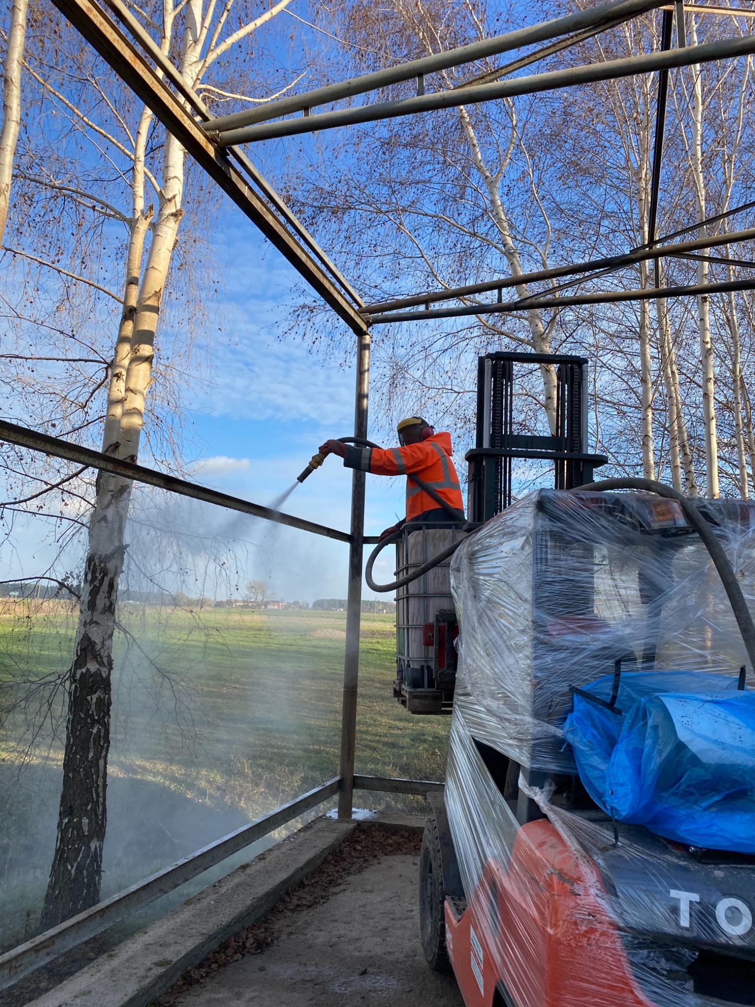 Pracownik w pomarańczowej kurtce roboczej, stojący na podnośniku widłowym, czyści konstrukcję metalową strumieniem wody pod ciśnieniem na tle drzew i pola.