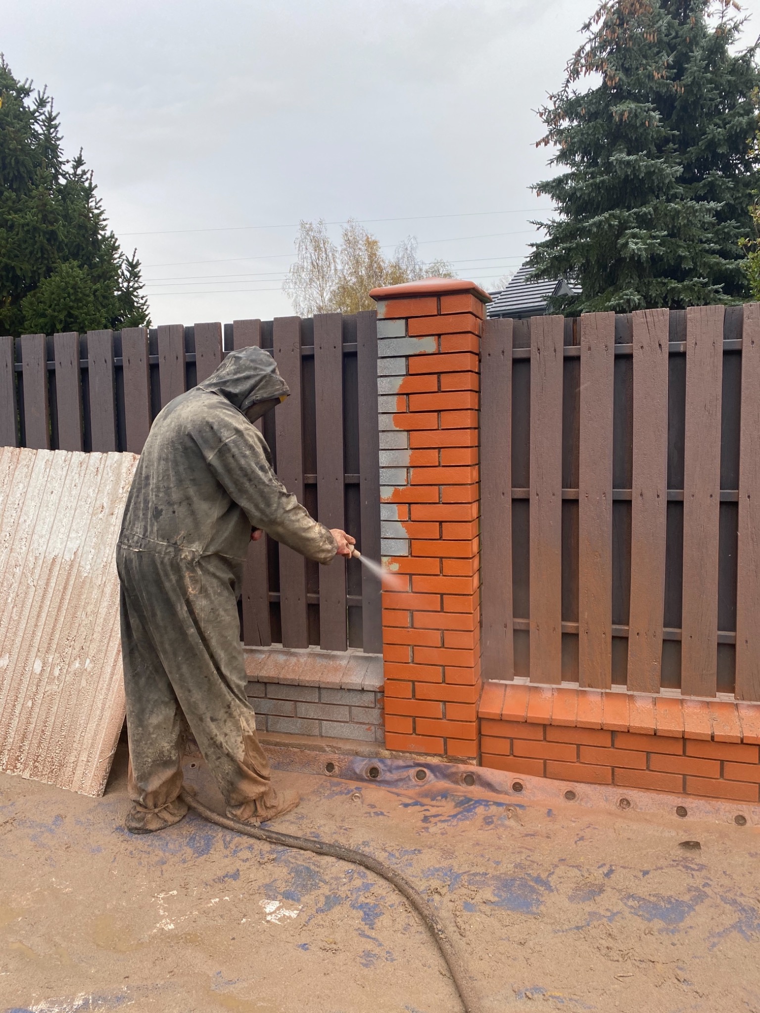 Pracownik w kombinezonie ochronnym nanosi powłokę na ceglany słupek ogrodzeniowy za pomocą pistoletu natryskowego; widoczne ślady zabrudzeń na podłożu.