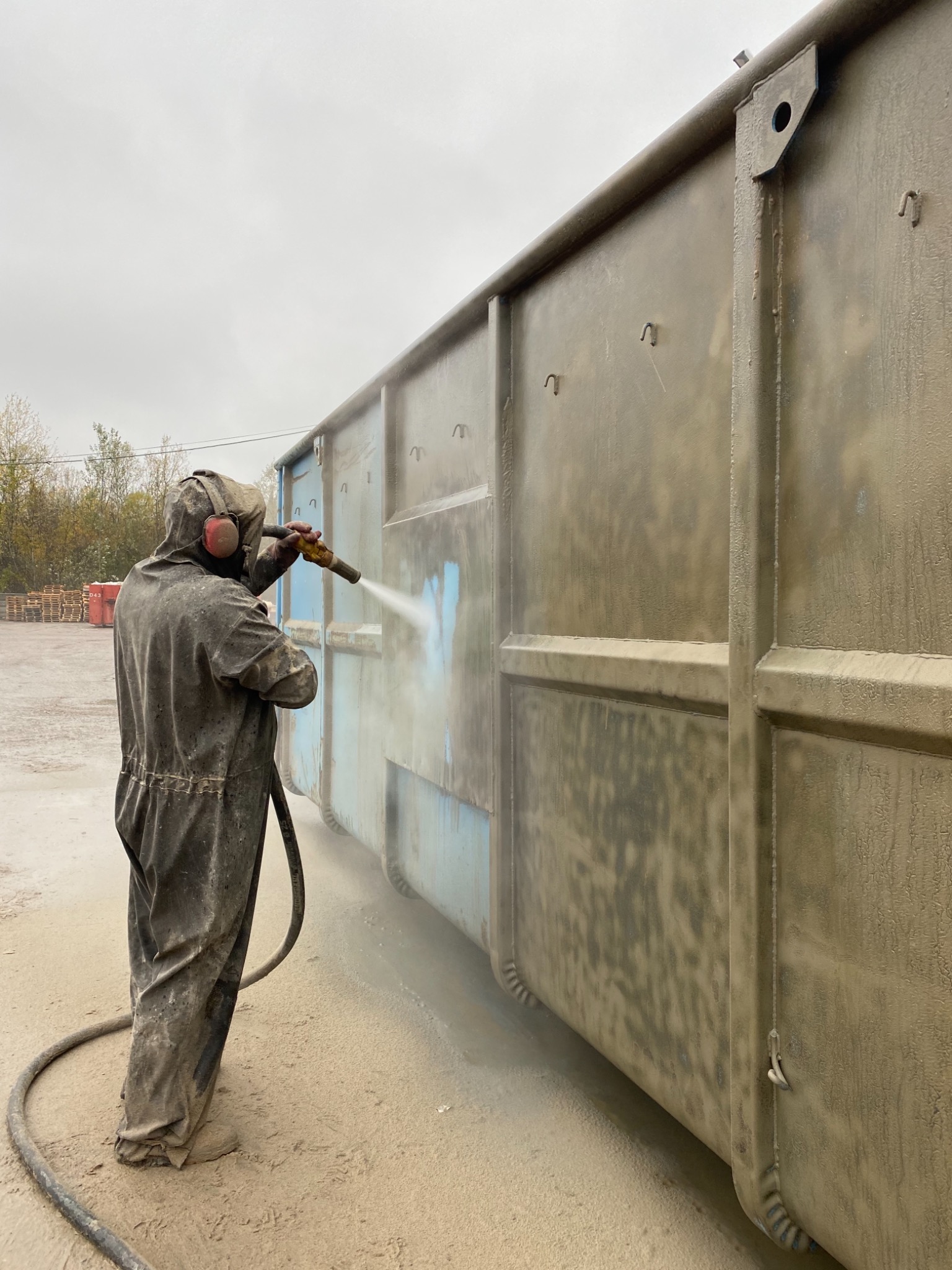 Pracownik w kombinezonie ochronnym piaskuje metalowy kontener na zewnątrz, widoczne ślady niebieskiej farby pod usuwaną warstwą.
