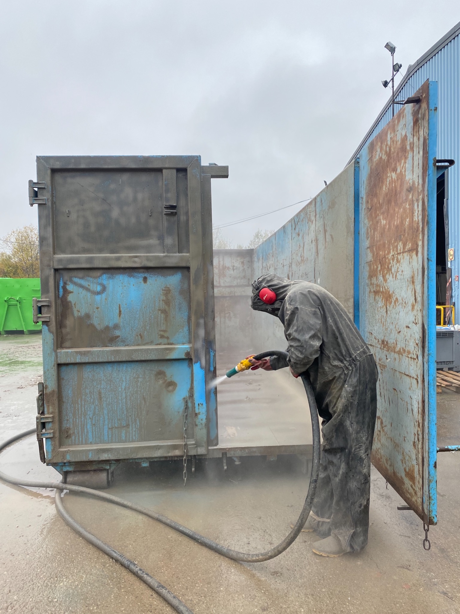 Pracownik w kombinezonie ochronnym czyści metalowy kontener myjką ciśnieniową, przygotowując go do dalszej obróbki.