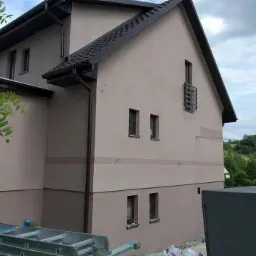 Dwukondygnacyjny dom z beżową elewacją, ciemnobrązowym dachem i rynnami, widoczny z boku, z drabiną opartą o ścianę i workami z materiałami budowlanymi w pobliżu.