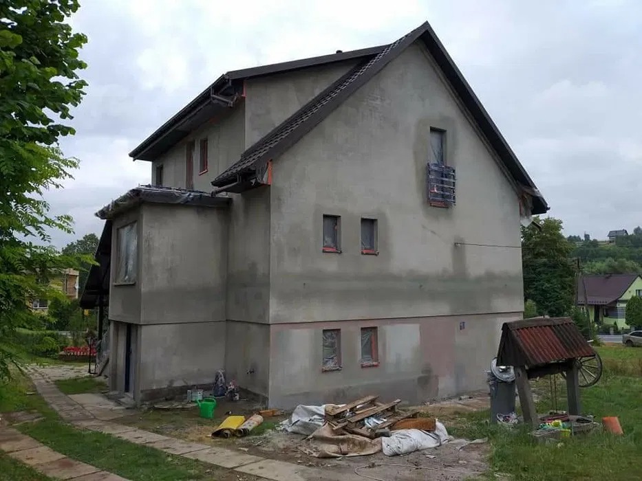 Dom w trakcie ocieplania elewacji, okna zabezpieczone folią, widoczne ślady świeżego tynku, w tle studnia z daszkiem i kołem.