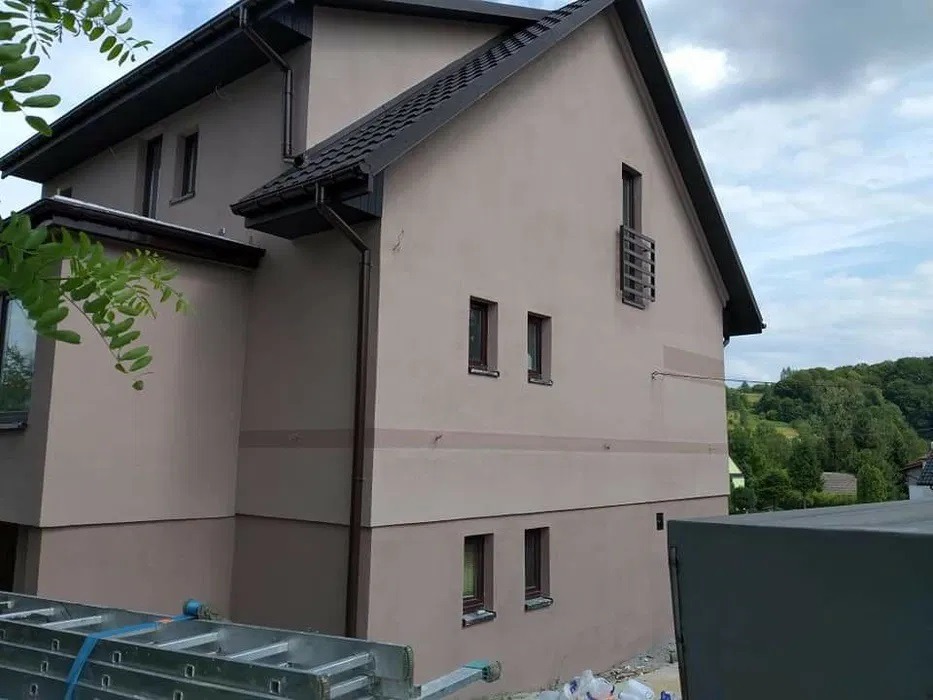 Dwukondygnacyjny dom z beżową elewacją, ciemnobrązowym dachem i rynnami, widoczny z boku, z drabiną opartą o ścianę i workami z materiałami budowlanymi w pobliżu.