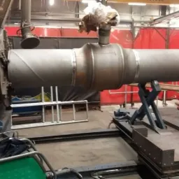 Metalowy element rurowy z widocznymi spawami, umieszczony na specjalistycznym stanowisku spawalniczym z automatycznym obrotnikiem, przygotowany do dalszej obróbki.