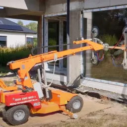 Pomarańczowy podnośnik próżniowy Smartlift montuje duże okno PCV w surowym budynku, widoczne detale mocowania i otoczenie budowy.