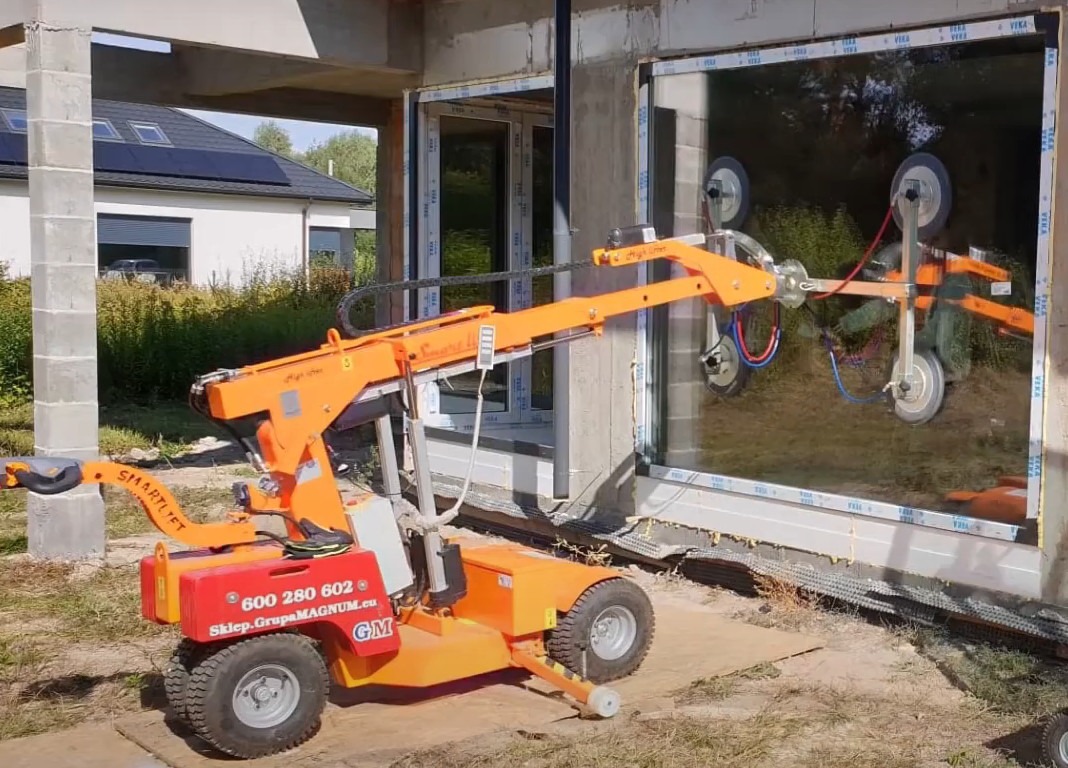 Pomarańczowy podnośnik próżniowy Smartlift montuje duże okno PCV w surowym budynku, widoczne detale mocowania i otoczenie budowy.