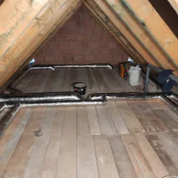 Instalacja przewodów wentylacyjnych w przestrzeni poddasza, widoczne drewniane krokwie, ocieplenie folią aluminiową i fragment ściany z cegły, na podłodze leżą różne przedmioty.
