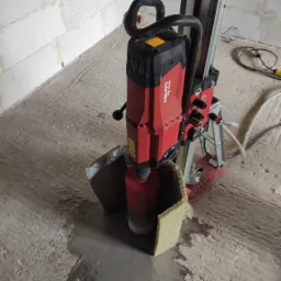 Czerwona wiertnica Hilti DD 250 z rdzeniem diamentowym wierci otwór w betonowej podłodze, otoczona osłoną z wełny mineralnej, widoczne mokre ślady po wierceniu.