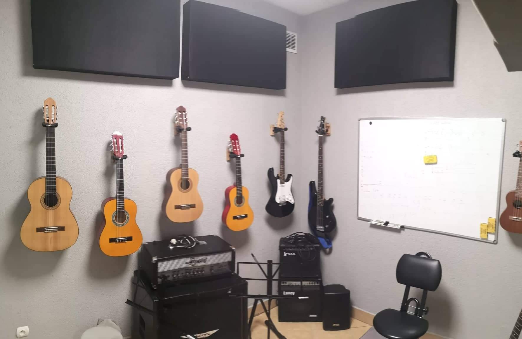 Salka z kilkoma gitarami klasycznymi i elektrycznymi wiszącymi na ścianie, wzmacniacze, tablica suchościeralna i panele akustyczne.