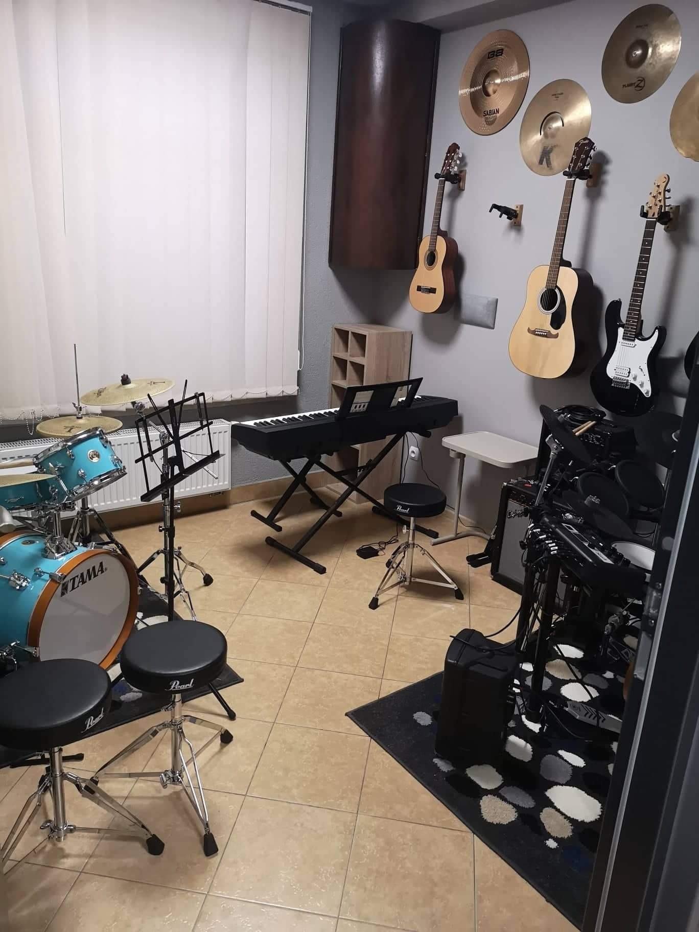 Salka prób z błękitną perkusją Tama, pianinem cyfrowym, gitarami akustycznymi i elektryczną na ścianie oraz dywanem w plamy.